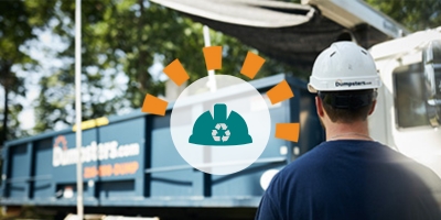Worker in a white hard hat standing in front of a white service truck hauling a Dumpsters.com roll off bin.