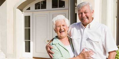 An elderly couple smile while standing outside of a home.