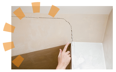 A hand pointing at a crack on a tan wall being held up by a white support beam. 