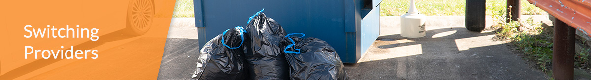 A silver car next to a blue front load dumpster with three garbage bags in front of them.