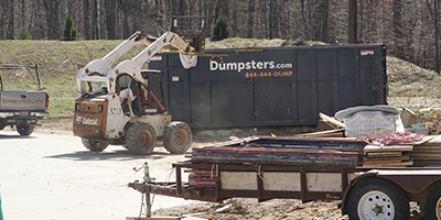 A skid steer loader emptying debris into a Dumpsters.com dumpster at a construction site.