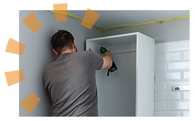 A man in a white shirt supports the bottom of a white kitchen cabinet while a man in a gray shirt unfastens it from the wall.