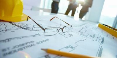 Workers talk in the background while glasses, a hard hat and a pencil sit on blueprints.
