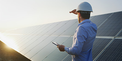 An EPC contractor wearing a white hard hat looking toward the sun next to a line of solar panels with a tablet in their hand.