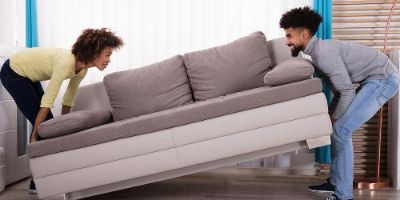 Young Man and Woman Smiling at Each Other While Bending Their Knees to Lift a Gray Couch