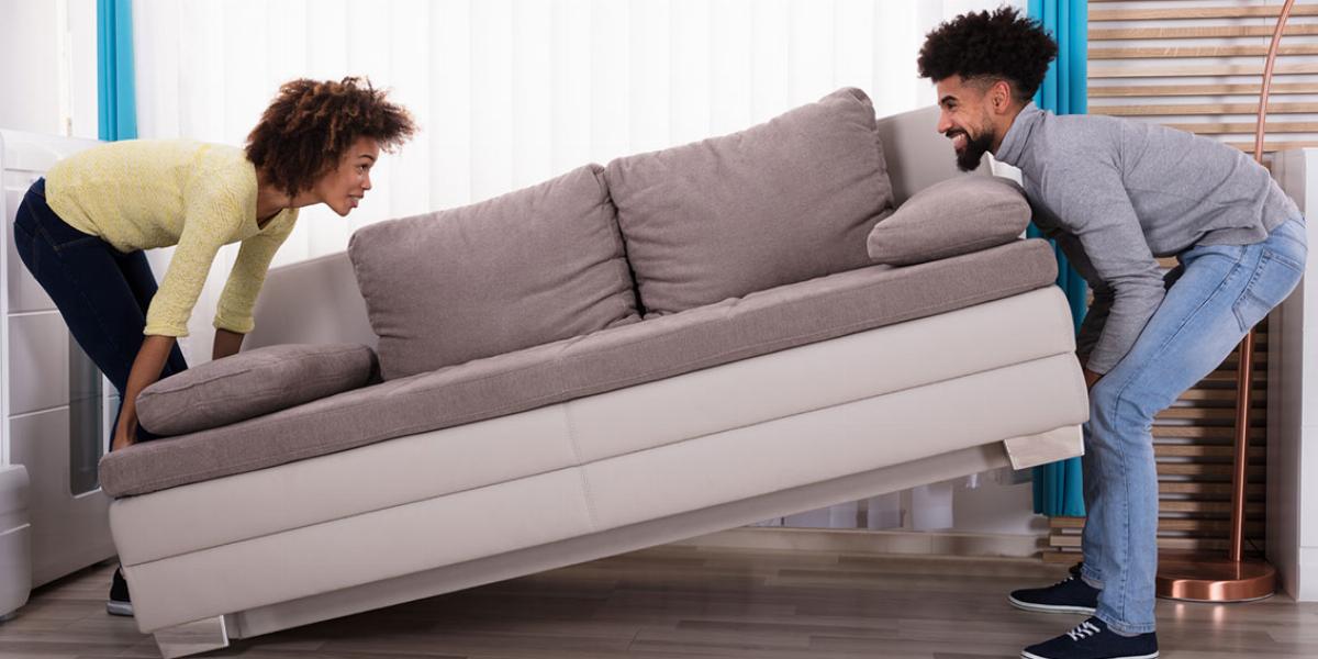 Young Man and Woman Smiling at Each Other While Bending Their Knees to Lift a Gray Couch