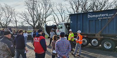 Construction worker in an orange vest and white helmet holding a cellphone to their ear on an active jobsite, with the Dumpsters.com orange radial arc overlaid.
