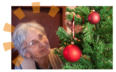 An older woman decorating an artificial Christmas tree with a red ornament.