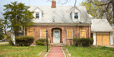 A foreclosed property with boarded up windows and doors and an overgrown yard.