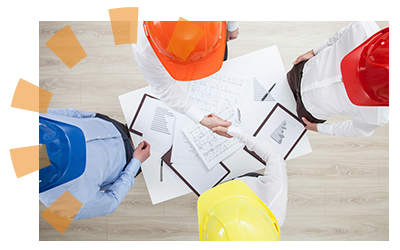 Four workers wearing brightly colored helmets shaking hands and standing over blueprints.