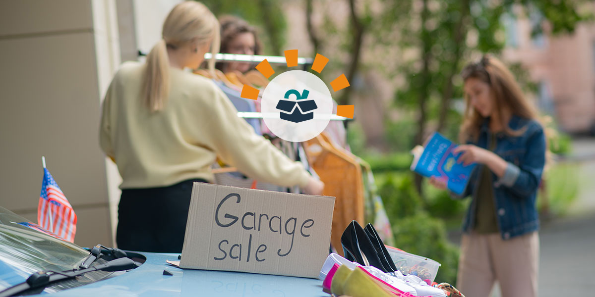 Garage sale box sitting on a blue car with clothing items on display and women shopping in the distance.