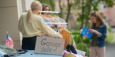 Garage sale box sitting on a blue car with clothing items on display and women shopping in the distance.