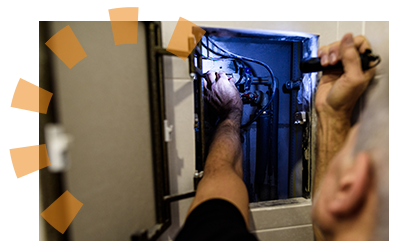 A man holding a flashlight looking at old plumbing and electric in a wall.