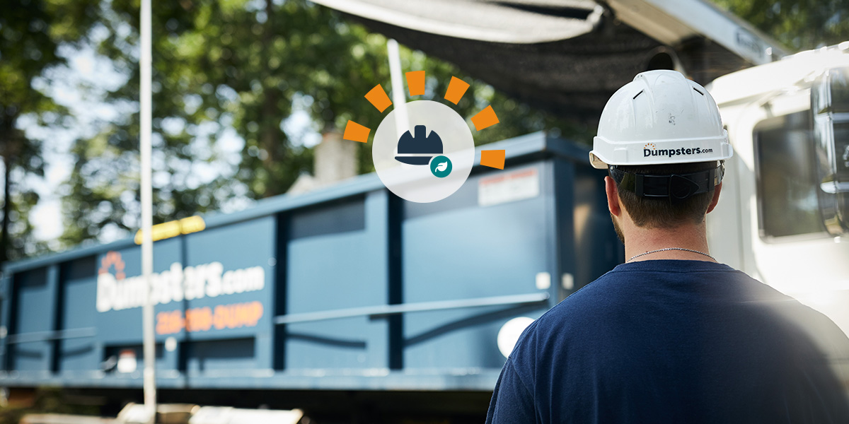 Worker in a white hard hat standing in front of a white service truck hauling a Dumpsters.com roll off bin.