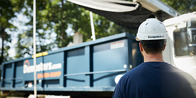Worker in a white hard hat standing in front of a white service truck hauling a Dumpsters.com roll off bin.