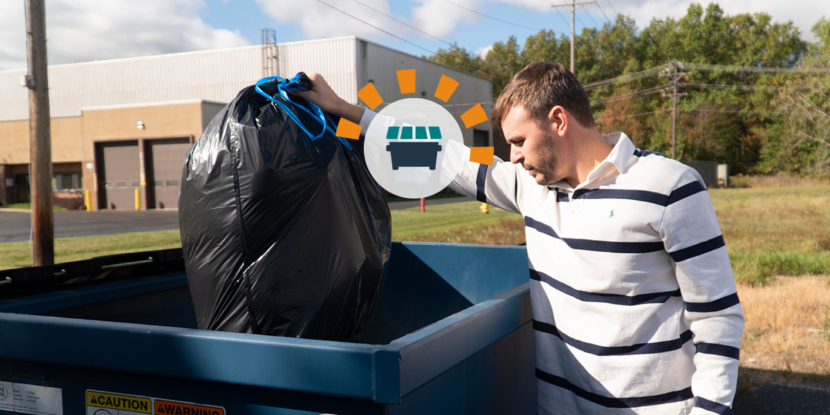 A man in a white shirt with black stripes placing a trash bag in a blue dumpster.