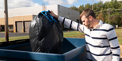 A man in a white shirt with black stripes placing a trash bag in a blue dumpster.