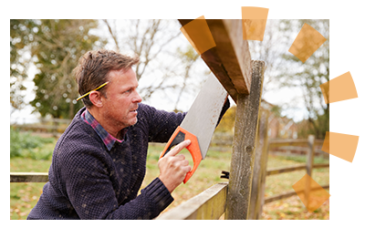 Male homeowner with purple sweater, pencil behind ear, hand sawing apart old fence.