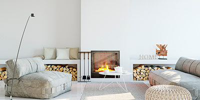 A white living room with a fireplace and firewood, grey couches and a white coffee table. 