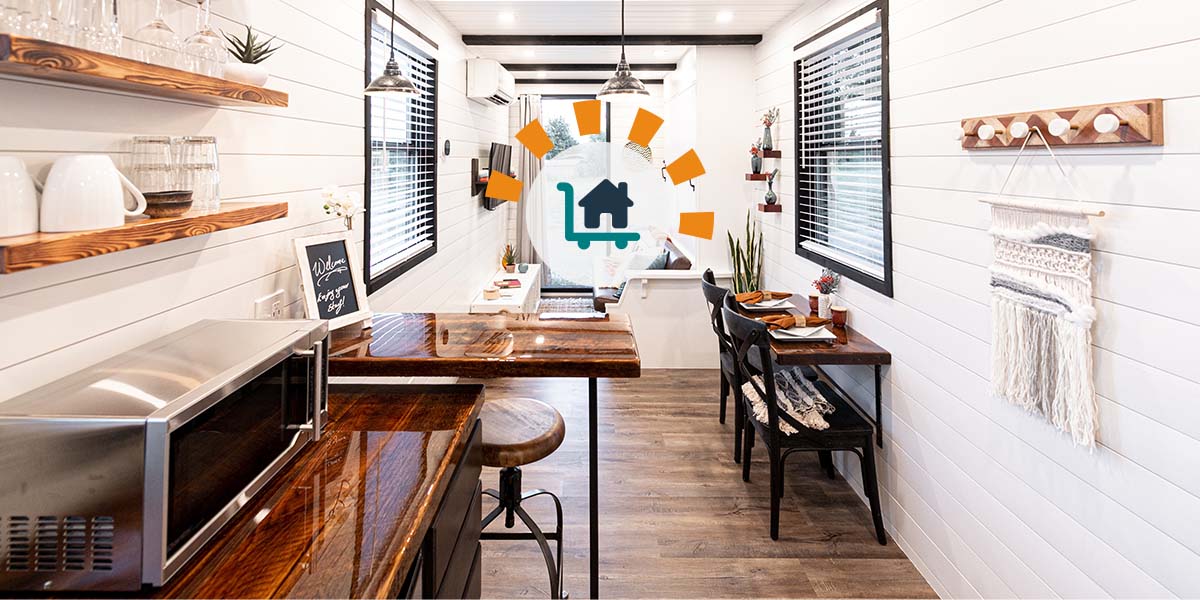 View from the kitchen of a modern tiny home with bright white shiplap walls, rustic wood countertops and shelves, and a sitting area on the other side of the small space.