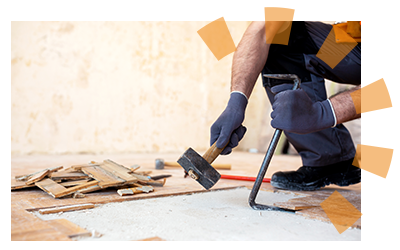 Close up view of a pry bar wedged beneath a piece of wood flooring and a mallet in motion to knock it loose with flooring debris in the background.