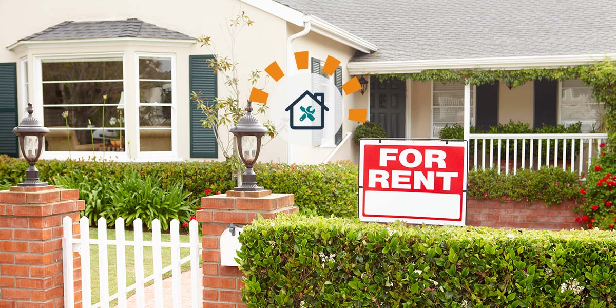 Red for rent sign on white wooden stake in front lawn of yellow multi-story house with front porch.