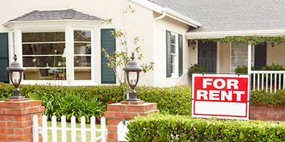 Red for rent sign on white wooden stake in front lawn of yellow multi-story house with front porch.