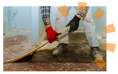 A man in work clothes and protective gloves tearing out old flooring exposing the subfloor.