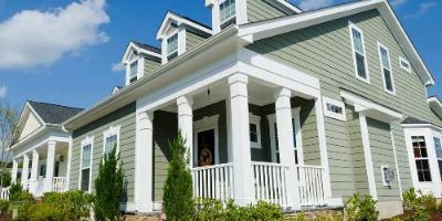 A gray home with white trim situated on a corner lot with green bushes and a blue sky