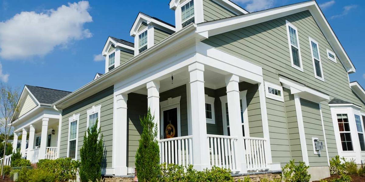 A gray home with white trim situated on a corner lot with green bushes and a blue sky