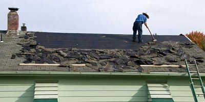 A man scrapes shingles off of the roof of a green house.
