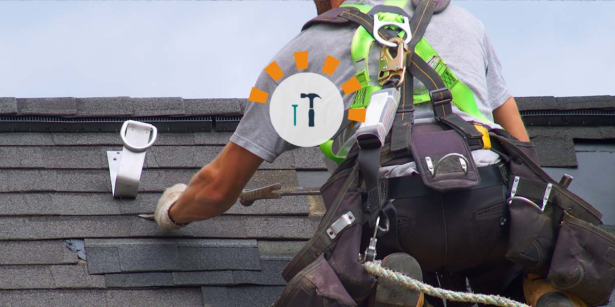 A roofer working on an asphalt shingled roof while attached to a safety harness.