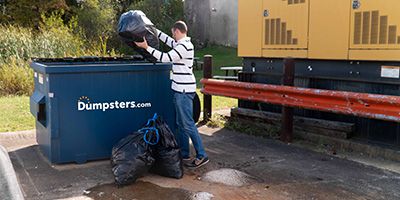 A man tossing a trash bag in a front load dumpster.