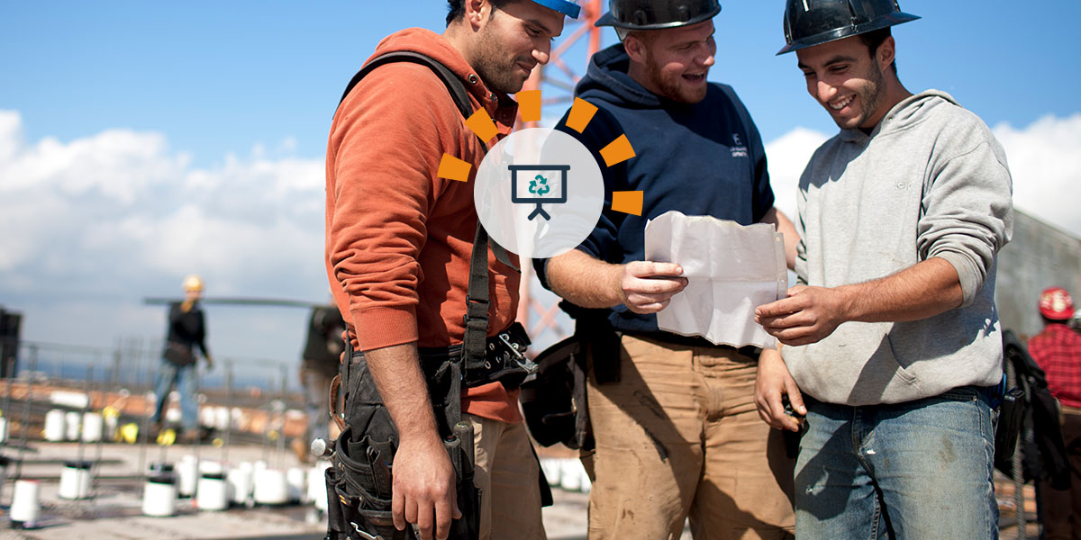 Three construction workers in hard hats kneeling on concrete at a jobsite and looking at construction plans.