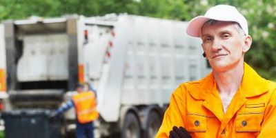 Garbageman in orange long-sleeve shirt with white hat and black gloves stands cross-armed in foreground as white garbage truck is loaded by another garbageman in the background.