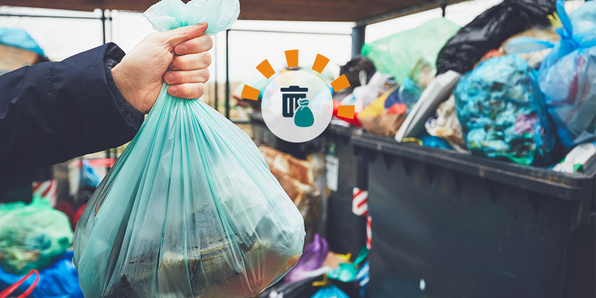 A hand holding a green trash bag in front of several overflowing trash bins. 