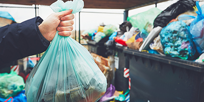 A hand holding a green trash bag in front of several overflowing trash bins.