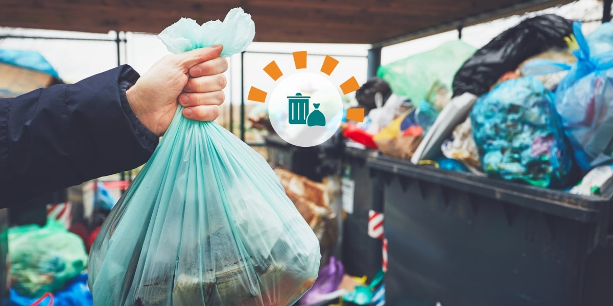 A hand holding a green trash bag in front of several overflowing trash bins.
