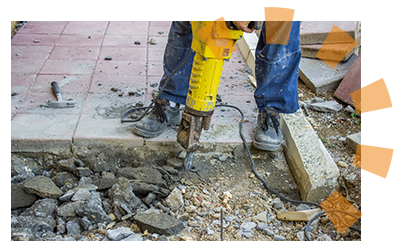 A man in work boots and jeans using a yellow jackhammer to break concrete. 