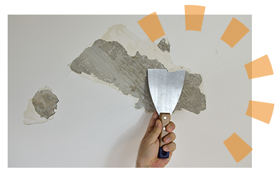 A hand holding a plaster knife removing plaster from a wall.