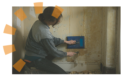 A woman kneeling near a wall using a steamer to scrape remaining wallpaper