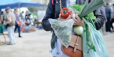 A customer holds vegetables and plastic grocery bags at a farmer’s market.