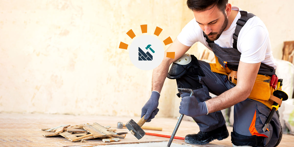 Young man using a pry bar and mallet to remove hard wood floors, wearing adequate safety gear like gloves and knee pads.