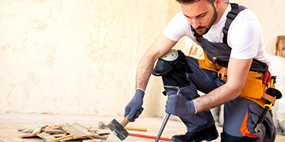 Young man using a pry bar and mallet to remove hard wood floors, wearing adequate safety gear like gloves and knee pads.