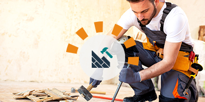 Young man using a pry bar and mallet to remove hard wood floors, wearing adequate safety gear like gloves and knee pads.