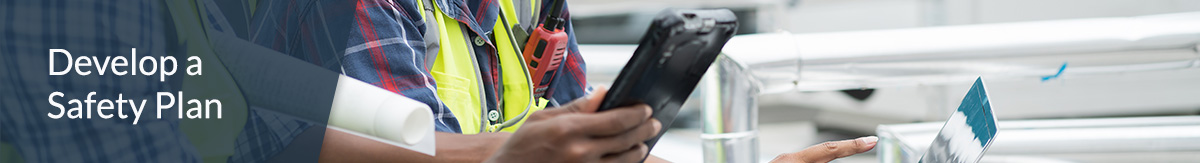 Two seated construction workers wearing white hard hats and yellow safety vests in discussion while using a laptop and tablet at a construction site. 