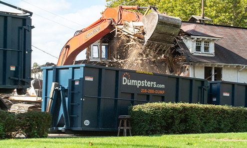 Crane Demolishing Home Behind Dumpsters.com Dumpster