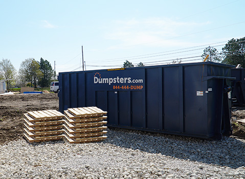 Blue Dumpsters.com dumpster sitting on gravel next to a stack of wood pallets.
