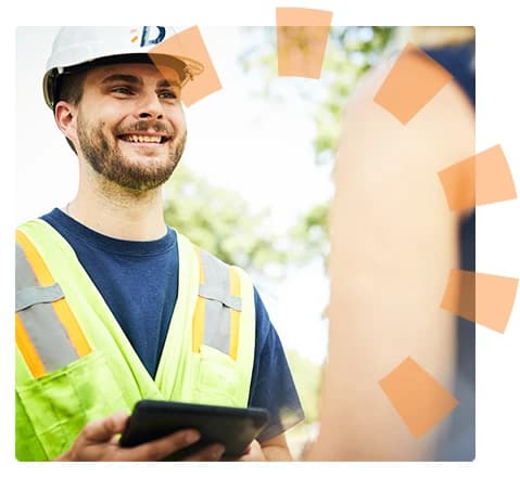 A smiling dumpster driver wearing a hardhat and safety vest is holding a tablet.