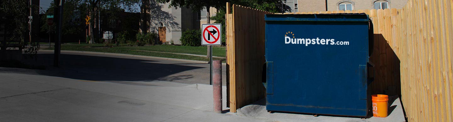 Dumpsters.com front load can sitting in a wooden dumpster enclosure in a parking lot.